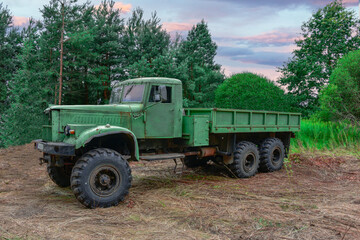 Stary, zdezelowany  samochód ciężarowy. Jest on pomalowany na zielony kolor i stoi w lesie na tle drzew. Niebo jest lekko zachmurzone. - obrazy, fototapety, plakaty