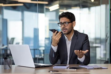 Upset and disappointed hispanic businessman recording audio message inside office at workplace, man complaining upset talking using smartphone app.