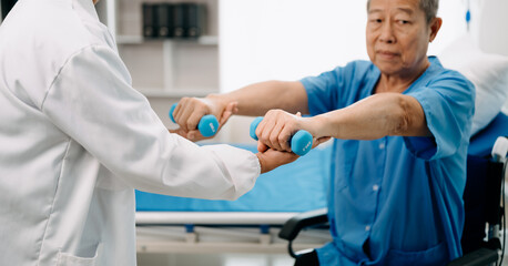 Asian physiotherapist helping elderly man patient stretching arm during exercise correct with dumbbell in hand during training hand in bed in clinic