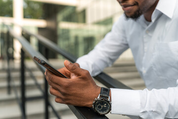 A portrait of a handsome Black businessman leaning against a railing during his break, holding a phone in his hand. 