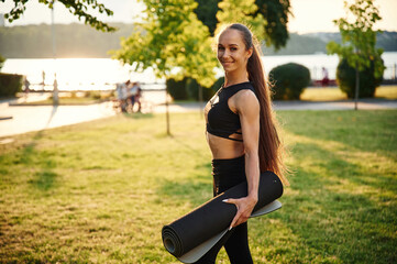 Obraz na płótnie Canvas Standing and holding yoga mat. Beautiful young woman in sportive clothes is in the park at sunny daytime