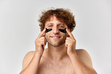 Fototapeta premium Portrait of mature shirtless man with curly hair taking care after skin, applying under eye patches over grey studio background. Concept of mens beauty, body and skin care, cosmetology, wellness, ad