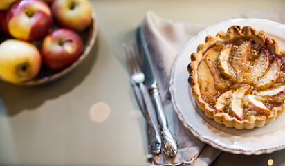 French Apple Tart baked with shortcrust pastry, also called Pâte Brisée, on table with cutlery and fresh apples, blurry bokeh lights for cozy Christmas holiday mood in background with copy space - 647589570