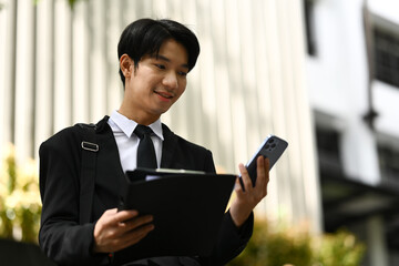 Fototapeta premium Young startup businessman using smartphone standing outdoors with office building in the background