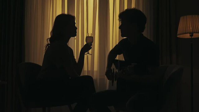 Everyday life creative concept. Young couple sitting in the living room, man playing the guitar, woman listening and drinks glass of wine.
