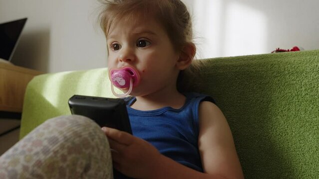 Cute caucasian baby girl sucking a pacifier while playing video game console. Computer entertainment.