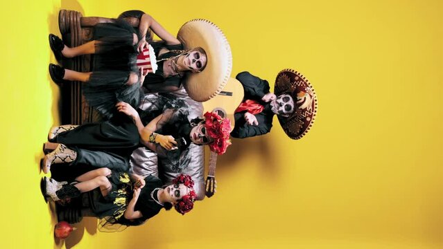 Man, woman and children in sugar skull makeup and costumes watching tv over yellow studio background. Concept of halloween, El Dia de Muertos, holidays and festivals, mexican traditions, family