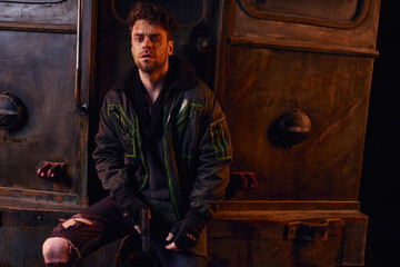 unshaven grim man with gun wearing worn clothes and looking at camera near rusty subway carriage