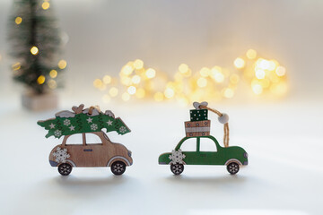Toy cars on a white background. A green wooden car with New Year's gifts on the roof and a beige car with a Christmas tree. Christmas tree and bokeh lights in the background.