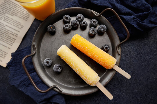 Overhead view of an ice lolly in a glass of orange juice with frozen blueberries