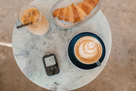 Close-up of a cup of coffee, iced coffee, croissant and glucose monitor on a table