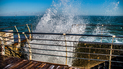 Large waves crashing on a wooden pier with railings against a blue sky - Powered by Adobe