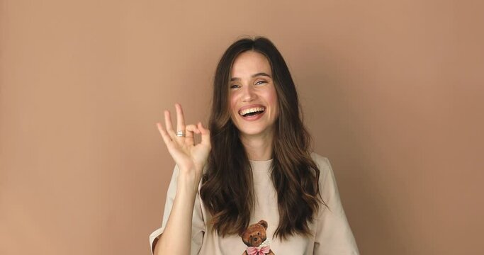 Okay! Playful attractive brunette woman showing ok and smiling, winking to camera, gesturing all right, agree sign, approving right choice isolated on beige background. All right!