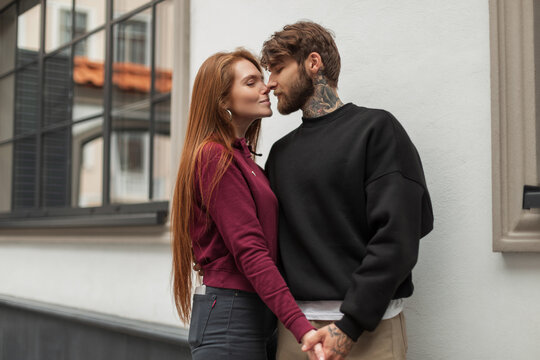Beautiful fashionable couple of lovers in fashion urban clothes kissing on the street. Handsome brutal man with a beard with a tattoo and a beautiful fresh red-haired girl in a hoodie outdoors