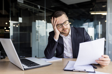 Young businessman sitting at desk in office and holding head, upset and worried reading documents,...
