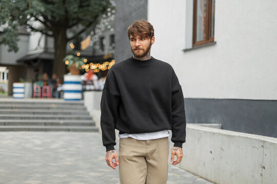 Fashionable handsome hipster man with a beard and tattoo in a black stylish mockup pullover walks in the city