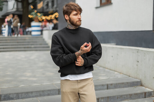 Handsome fashionable brutal hipster man with a tattoo in a fashion black pullover walks in the city
