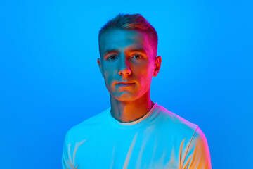 Portrait of young good-natured, calm man, student in white t-shirt looking at camera isolated on blue background in red neon light.