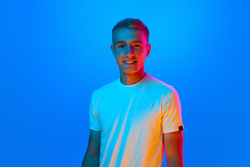 Portrait of young happy, smiling man, student in white t-shirt looking at camera isolated on blue background in red neon light, filter.