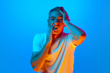 Portrait of young man, student on blue studio background in neon light, tired covering face hands, depression and sadness, upset and irritated for problem