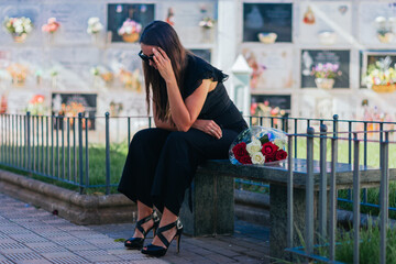 Weeping woman with sunglasses, black dress and hand on her head, sitting on a bench, with a bouquet...