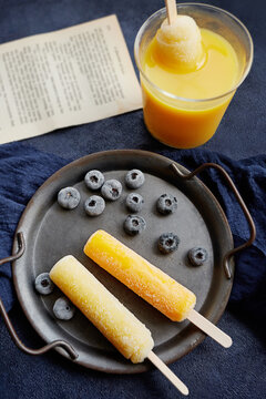 Overhead view of an ice lolly in a glass of orange juice with frozen blueberries