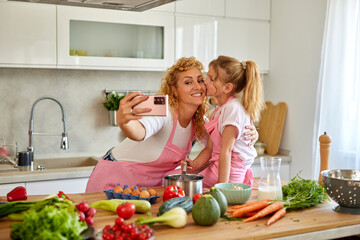 Beautiful woman and her cute daughter taking selfie in the kitchen while preparing food
