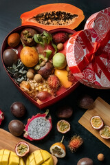 An assortment of fresh exotic fruits arranged on blacktop, wooden boards. Some of the fruits are cut and peeled. Some of the fruits are in a gift box