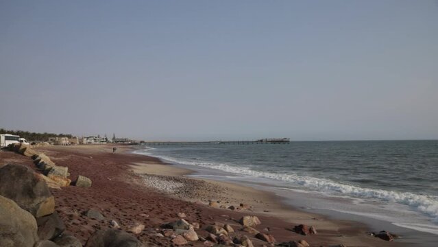 A beach with red, black and white sand and multiple types of rocks with the Swakopmund jetty in the background