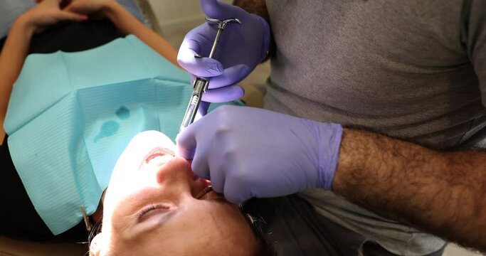 Dentist in blue gloves doing painkiller injection for woman during wisdom tooth removal. Doctor works with woman sore dental spots slow motion