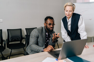 Obraz premium Focused diverse business partners looking at laptop screen, discussing corporate software application. Young black male boss leader consulting about project with female intern employee.
