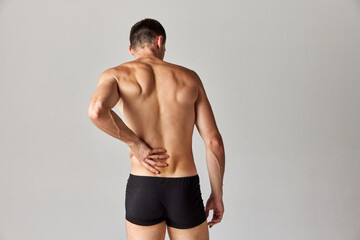 Back pains. Muscular man standing in underwear and holding his back against grey studio background....