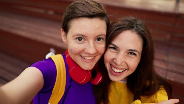 Portrait of a brunette girl in a yellow sweater and a girl with a short haircut in a purple top and red headphones who take a selfie on a brown bench in a skatepark in summer