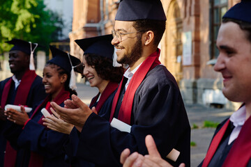 Fototapeta premium Graduated students clapping hands while standing outdoors near the university