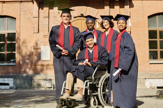 Portrait of happy group of students with their classmate with disability smiling at camera while standing outdoors, they graduating from university