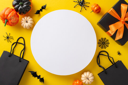 Get into the Halloween spirit with our sale: Top shot of black paper bags, giftbox with bow, pumpkins, spiders, cobweb, bats on yellow backdrop. Circular frame with ad space available