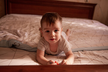 Fototapeta premium One year old baby lies on his stomach on the bed in the morning in the sun. The child is funny, smiling, playing in a great mood. Care, furniture for infants. Close-up portrait of a boy. Lifestyle