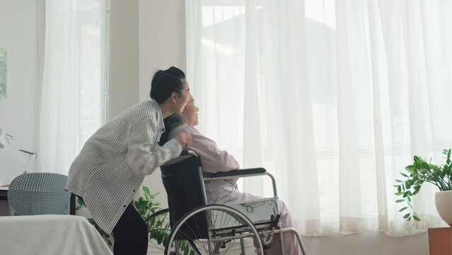 Medium long shot of young daughter pushing wheelchair of her sick senior mother to window in nursing home