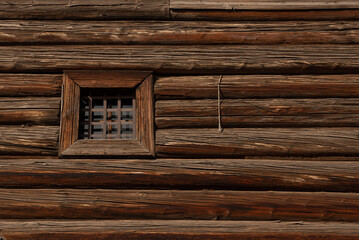 The wall of an old log house with a small window behind bars