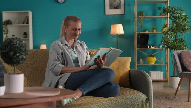 A woman reads a book with a smile, touched by the plot. A woman with a dreamy expression closes a book. A woman in casual clothes spends her day reading a book while sitting on the sofa at home.