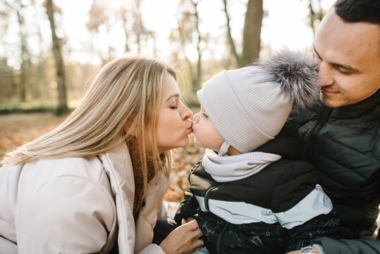 Mom kissing child, father, kid have picnic in autumn forest in nature. Family holiday, spending time together at sunset. Mother, dad hugs son sitting on blanket on golden leaves in park. Closeup