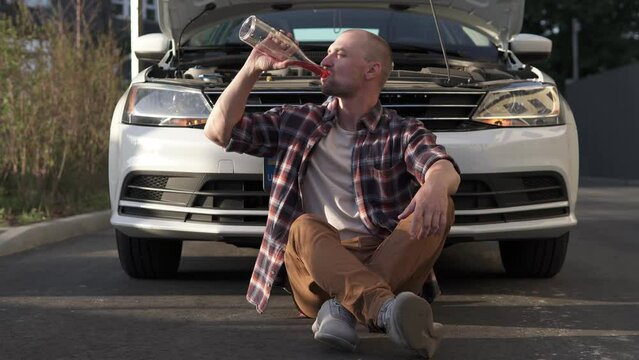 A male driver drinks alcohol near a broken car after an accident. Drunk driving concept.