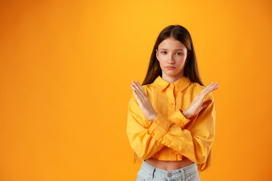 Teen Girl Shows Cross Hands Gesture, Rejecting Something Over Yellow Background In Studio