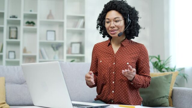 Confident african american businesswoman in wireless headset talking on video call using laptop while sitting in home office. Black woman coach conducts remotely an online training, seminar, webinar 