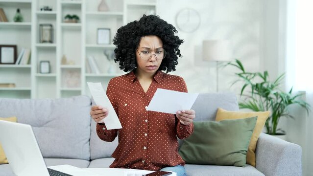 Frustrated african american female looking at large utility bills sitting on sofa at home. Upset black woman holding a paycheck, thinking about problems in the household budget and financial expenses