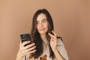 Astonished surprised brown haired girl wear casual T-shirt, using mobile phone for checking social networks, looking at camera with big eyes, standing isolated over beige background.