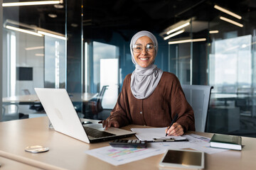 Portrait of young successful Arab woman in hijab working in office with documents and laptop. Smiling and looking at the camera.