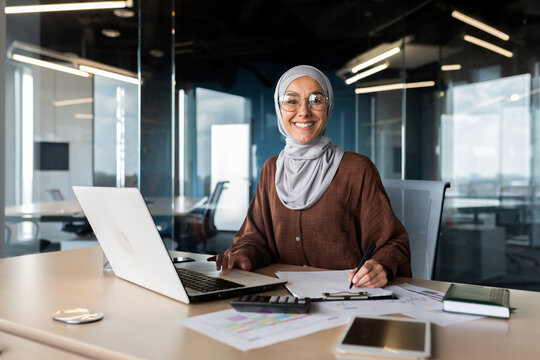Portrait of young successful Arab woman in hijab working in office with documents and laptop. Smiling and looking at the camera. - Powered by Adobe