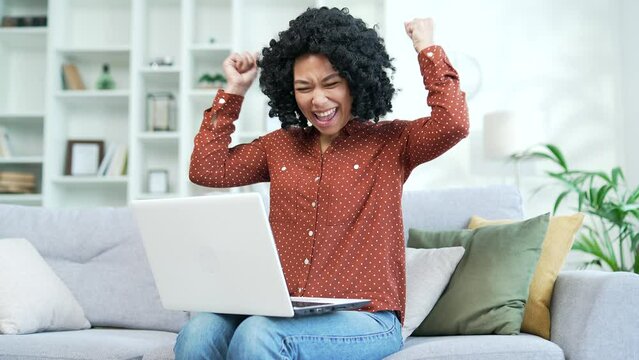 Happy young african american female celebrating success by reading great news on laptop computer in living room at home. Excited smiling black woman satisfied with pleasant message sitting on the sofa
