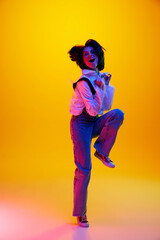 Full-length image of woman in her 30s, wearing casual clothes and cheerfully jumping over orange studio background in neon light. Concept of human emotions, fashion, lifestyle, facial expression, ad
